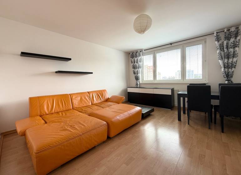 Priestranný 3-izbový byt s balkónom 68 m² Belinského - Obývačka v 3-izbovom byte s oranžovou sedačkou, čiernymi policami a jedálenským stolom.