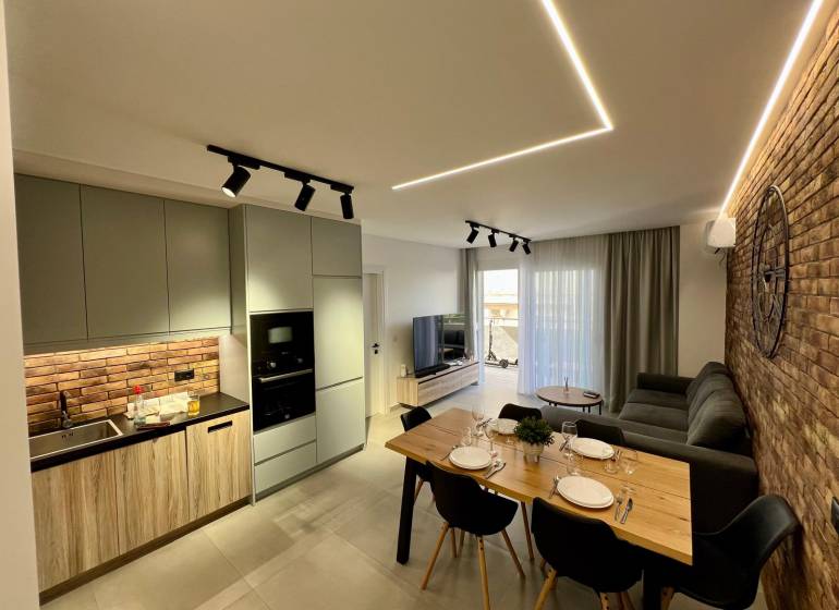 Sarandë - zariadený apartmán, výhľad na more, garážové státie - Obývačka s kuchyňou v Rekreačnom apartmáne. Tehlová stena, jedálenský stôl a pohodlná sedačka.