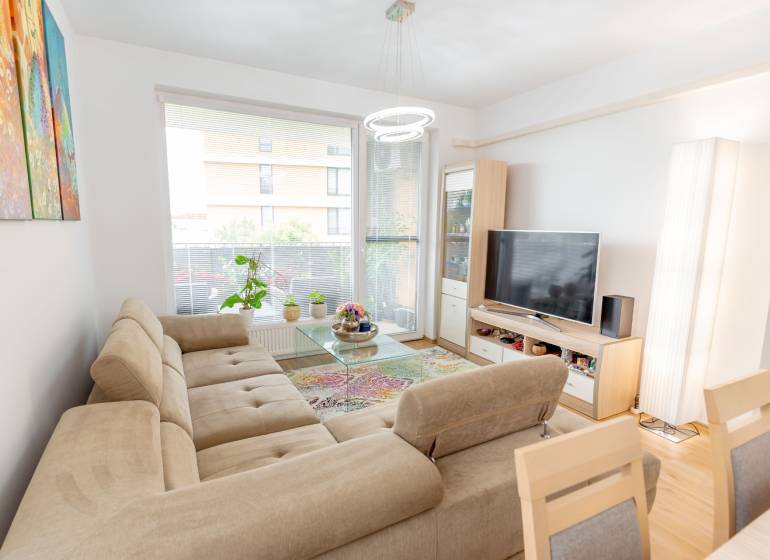 Jedinečný 3-izbový apartmán s balkónom, 71 m², Nový Ružinov - Svetlá obývačka 3-izbového bytu s pohovkou, televízorom, kobercom a kvetmi pri okne.