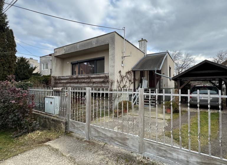 NA PREDAJ rodinný dom v Jelšovciach - EXKLUZÍVNE - Rodinný dom so záhradou, oplotením, dreveným prístreškom a bránou, v pochmúrnom počasí.