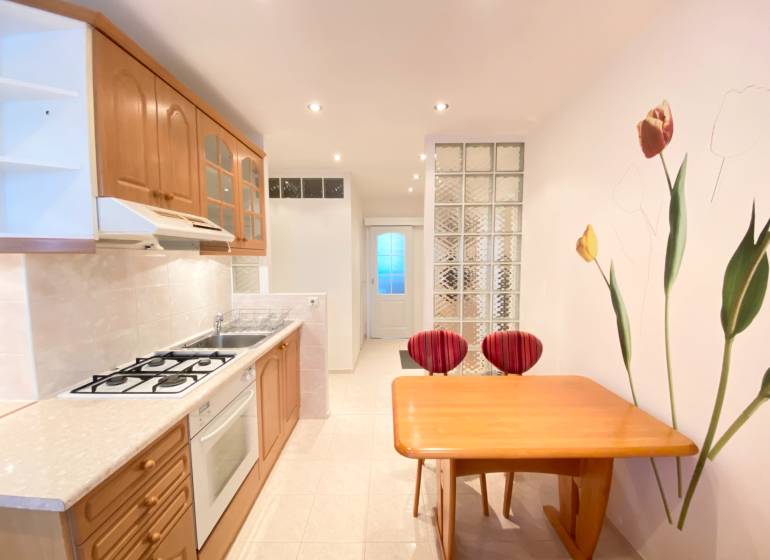 NA PRENÁJOM PRIESTRANNÝ 3-IZBOVÝ BYT V SKVELEJ LOKALITE, UL. SLOBODY - Kuchyňa v 3-izbovom byte s drevenými skrinkami, stolom a dekoratívnou stenou s kvetmi.