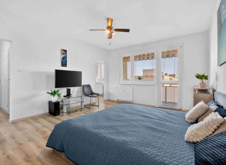 REZERVOVANÉ Predaj moderný 1-izbový byt, 45,24m2, Bratislava IV - Spálňa s posteľou, televízorom, kreslom, rastlinou a podlahou s dreveným dekorom v 1-izbovom byte.