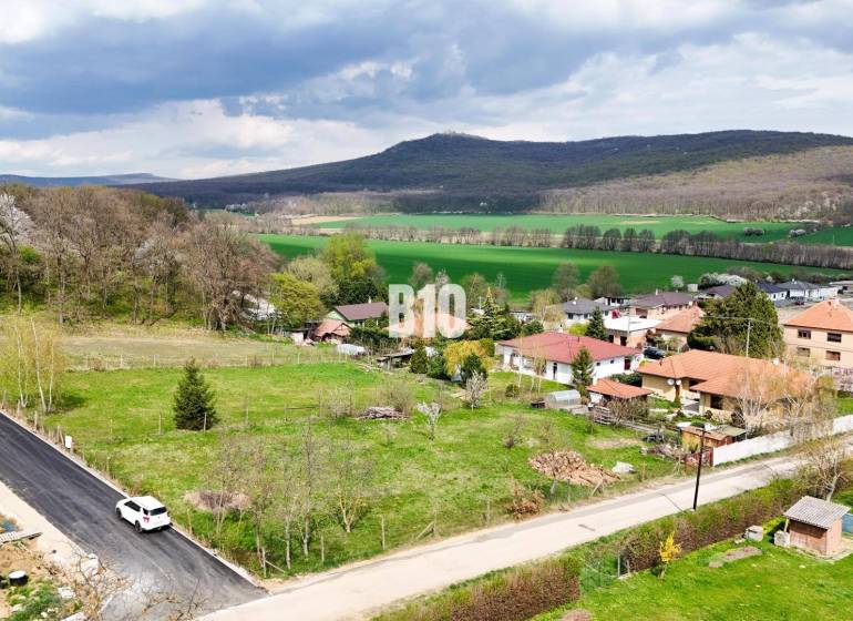 Jelenec  - Nádherný pozemok pri lese - 687 m2 - Pozemky - bývanie v Jelenci obklopené prírodou a rodinnými domami, s lesnatými kopcami v pozadí.