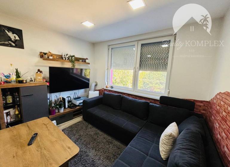 Vám ponúka exkluzívne na predaj moderný, komple - Obývacia izba s tmavou sedačkou, televízorom na stene a veľkým oknom.