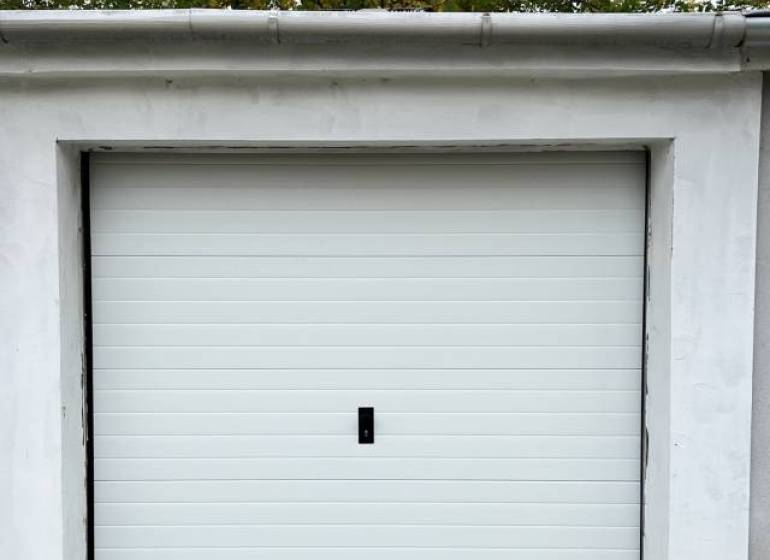 Vám ponúka na PREDAJ zrekonštruovanú garáž v Du - Garáže na Nevädzovej ulici v Dunajskej Strede s bielymi dverami a stromami v pozadí.