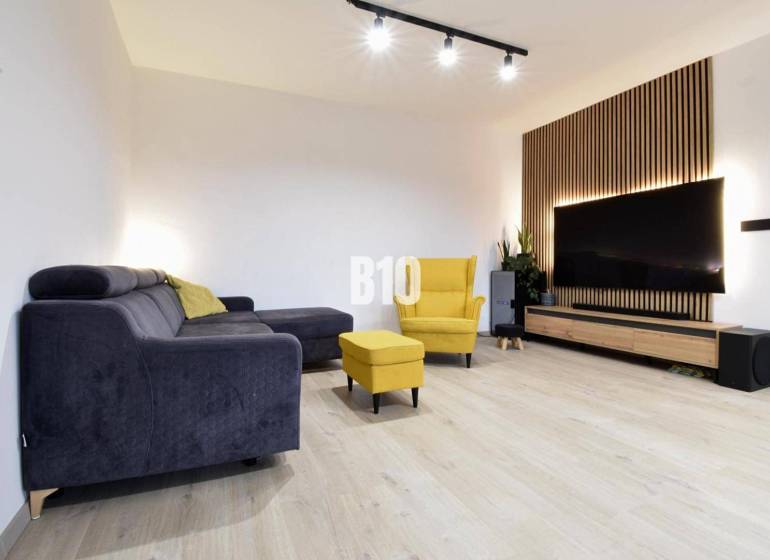 Veľkometrážny 3 izbový byt s garážou - Obývacia izba v 3-izbovom byte s podlahou s dreveným dekorom, TV a nábytkom.