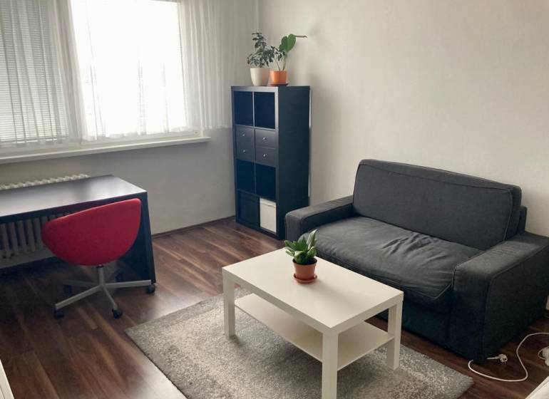 NA PRENÁJOM ÚTULNÝ 1-IZBOVÝ BYT, ČIASTOČNÁ REK., KAROLA ADLERA, BA - 1-izbový byt s pohovkou, stolom, červenou stoličkou, poličkou a podlahou s dreveným dekorom.