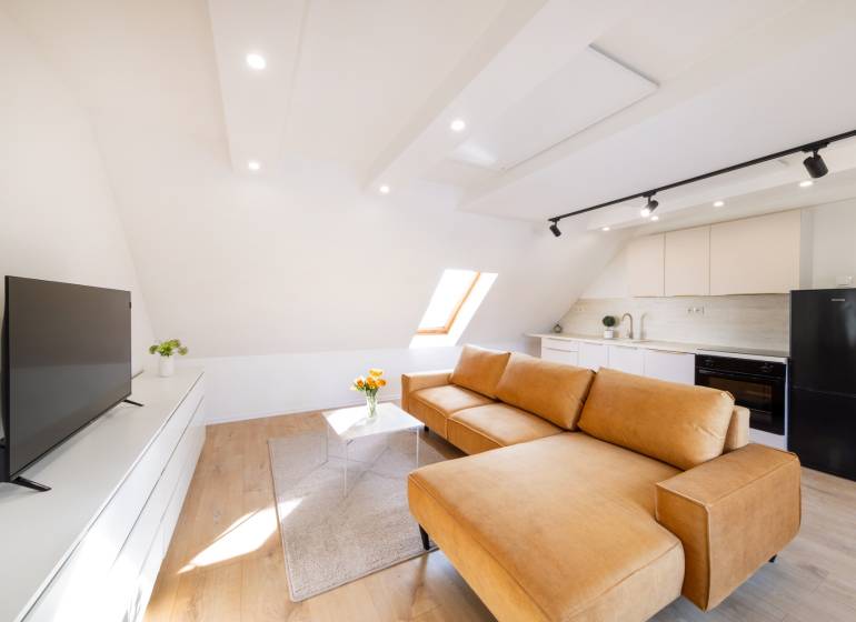  REZERVOVANY-PRENAJOM | NOVÝ 2 i. podkrovný byt v novostavbe - Obývacia izba s kuchyňou v 2-izbovom byte: veľké okno, hnedá sedačka, podlaha s dreveným dekorom.