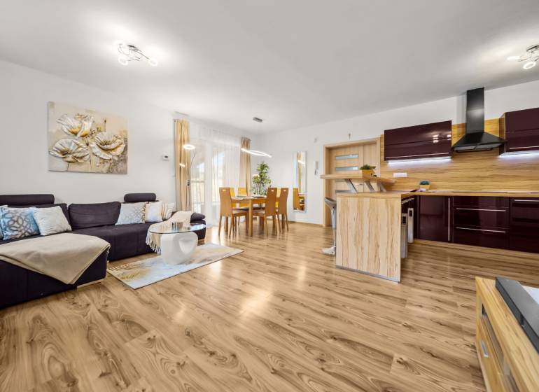 MODERNÝ 5-IZBOVÝ RODINNÝ DOM V ČUNOVE: 180m2 KOMFORTU A TICHA - Obývacia izba a kuchyňa v rodinnom dome s podlahou s dreveným dekorom, pohodlný gauč.