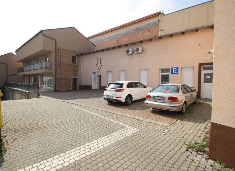 Ponúka na predaj budovu - centrum Senca - NOVÁ SUPER CENA  - Na ulici Mierové nám. pred budovou sú dve autá na parkovisku a smetné koše.