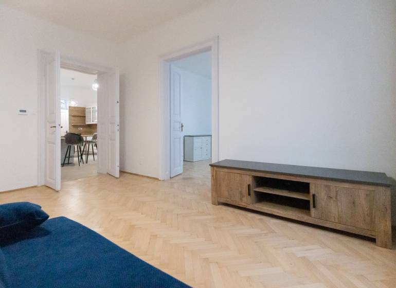 Priestranný 3i byt s vysokými stropmi, Dunajská - Obývacia izba 3-izbového bytu s modrou sedačkou a podlahou s dreveným dekorom.