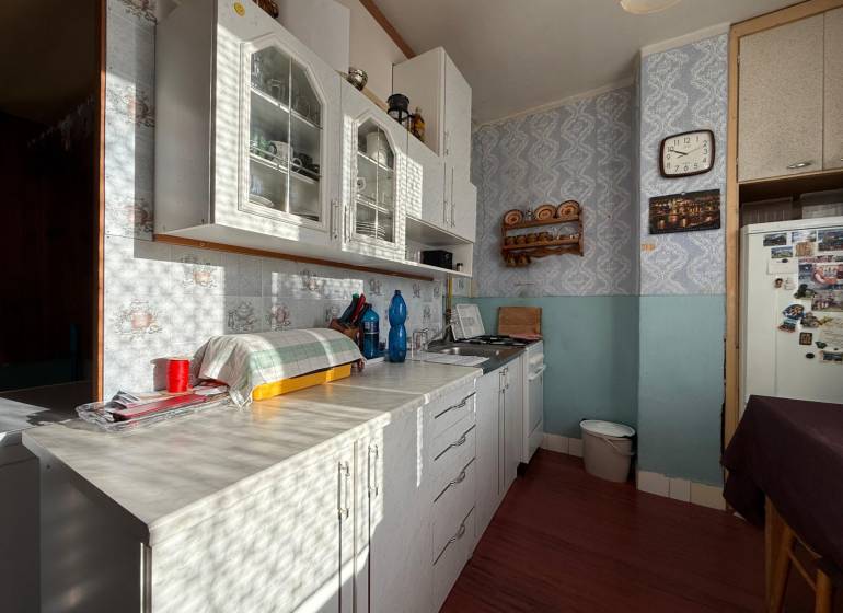 PREDAJ 2 IZBOVÝ BYT HANDLOVÁ  - Kuchyňa 2-izbového bytu so svetlou linkou, poličkou a retro hodinami na stene.