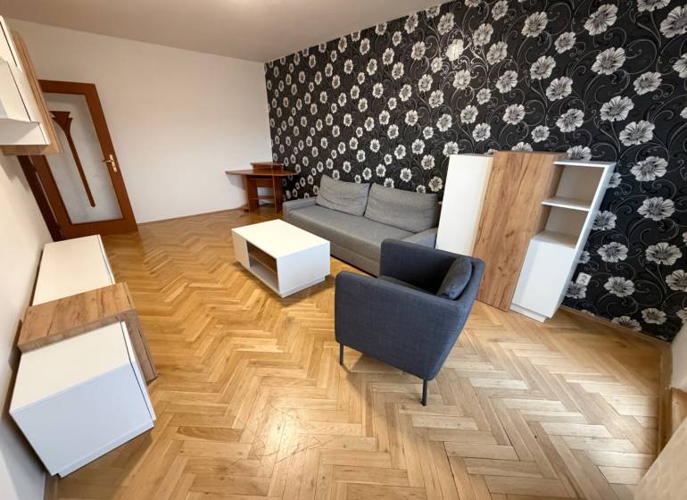 2 izbový byt s balkónom Topoľčany  - Obývačka v 3-izbovom byte s podlahou s dreveným dekorom, vzorovanou tapetou a nábytkom.
