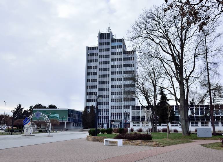 Kancelária s výhľadom na centrum Michaloviec. 16 m2 - Kancelárie na Námestí slobody v Michalovciach, vysoká budova s oknami a stromami v okolí.
