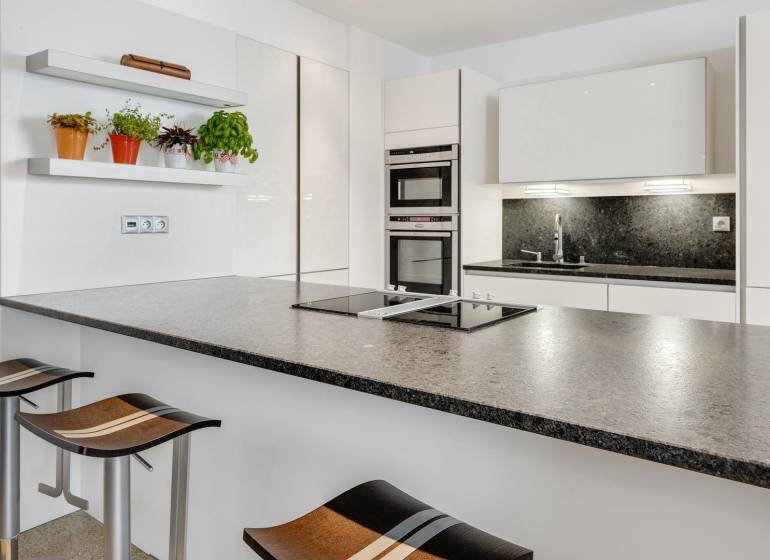 Novostavba 3 izbový byt, Palisády, 147 m2, výhľad, parkovanie, BA I - Kuchyňa v 3-izbovom byte s moderným ostrým a stoličkami, poličky s rastlinami.