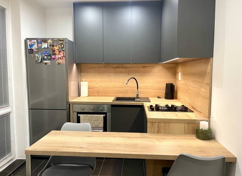 | Predaj 1-izbového bytu – Račianska, Bratislava - Kuchyňa v 1-izbovom byte s tmavými skrinkami a dreveným dekorom na doskách.