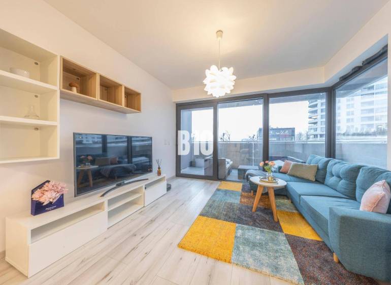 TEHELNÉ POLE | 1-izbový byt s priestrannou terasou - Obývacia izba v 1-izbovom byte s veľkou pohovkou, televízorom a podlahou s dreveným dekorom.