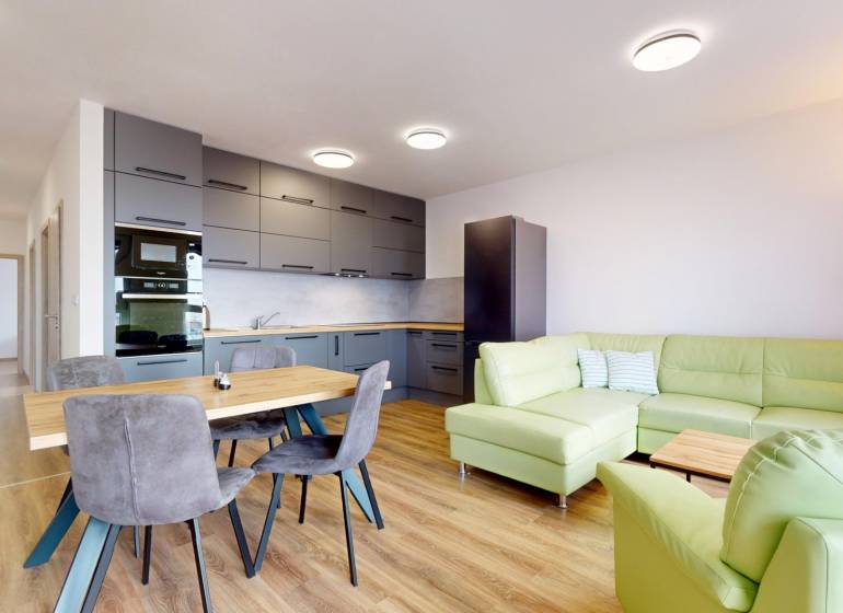 | 3-izbový byt v novostavbe, ul. Odevná + parking | Trenčín  - Kuchyňa s obývačkou v 3-izbovom byte, zelená sedačka, podlaha s dreveným dekorom.