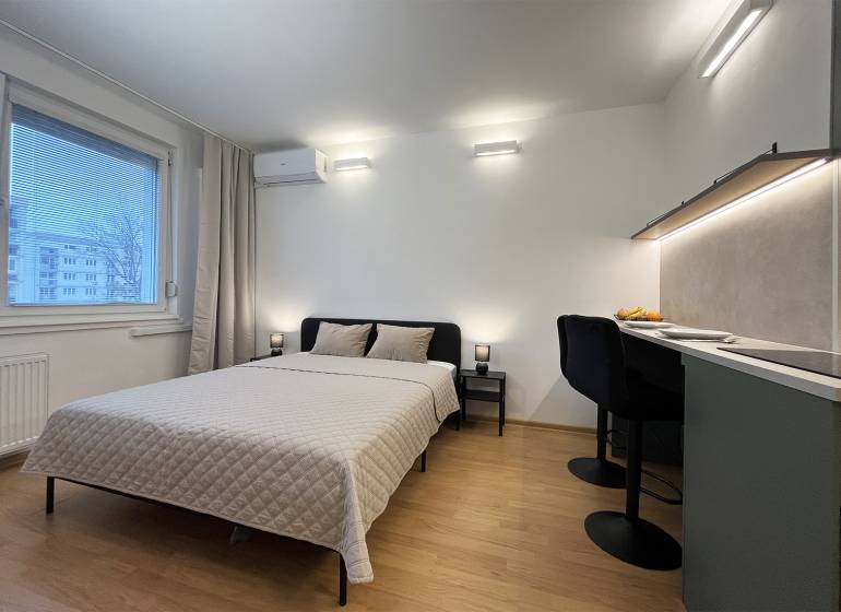 Bez provízie | Na prenájom dizajnové štúdio v Ružinove - Garsónka s posteľou, stolom a podlahou s dreveným dekorom, klíma na stene.