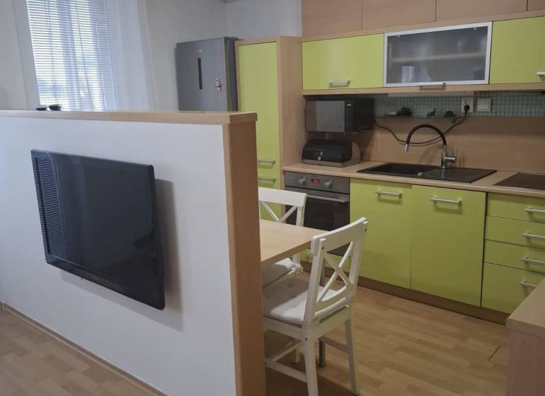 Na prenájom klimatizovaný 2 izbový byt v Moste pri Bratislave - Kuchyňa v 2-izbovom byte so zelenými skrinkami, televízorom a stolom s dvomi stoličkami.