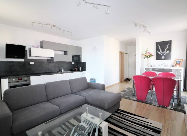 NUERA︱ SLNEČNICE- slnečný 2izbový byt s výhľadom na hrad - Obývacia izba s kuchynským kútom, sivou sedačkou, skleneným stolom a červenými stoličkami.