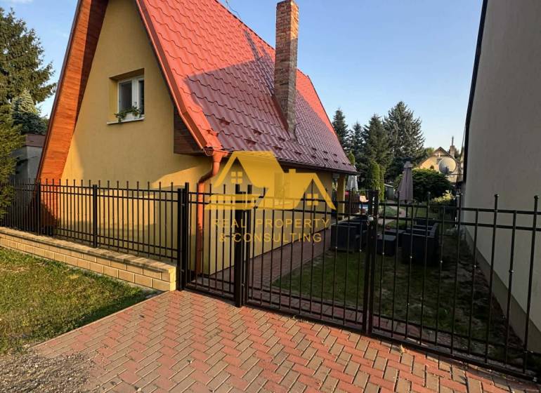 Predám peknú chatu na celoročné bývanie v Nových zámkoch - Chata na ulici v Nových Zámkoch, so špicatou strechou a tehlovým komínom.