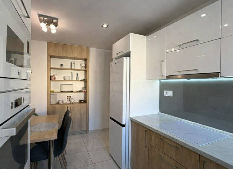 NA PREDAJ vkusne zrekonštruovaný  2 izbový byt, Komárno - Kuchyňa v 2-izbovom byte s bielymi skrinkami, stolom a dreveným dekorom políc.