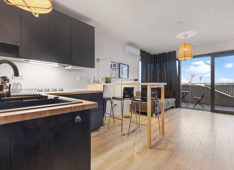 - PREDAJ - APARTMÁN - 38 m2 + 14 m2 LOGGIA - TEHELNÉ POLE - Kuchyňa s čiernymi skrinkami, barovým pultom a podlahou s dreveným dekorom v 1-izbovom byte.