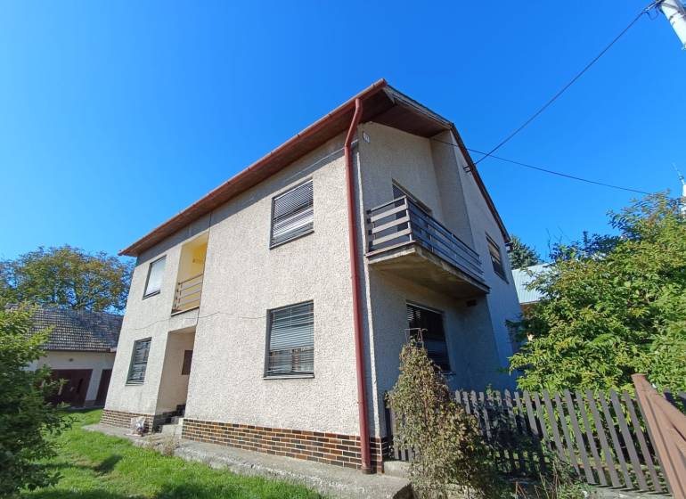 : Predaj rodinného domu s veľkým pozemkom v obci Pitelová  - Rodinný dom v Pitelovej s balkónom a záhradou, modrá obloha, plot a zeleň.