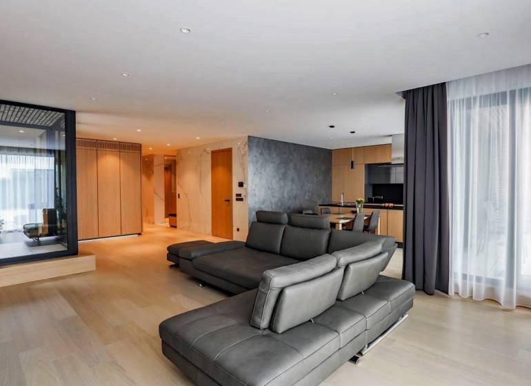RIVERS I Prémiový 4-izbový byt s panoramatickým výhľadom na mesto - Obývacia izba v 4-izbovom byte s podlahou s dreveným dekorom a koženou sedačkou.