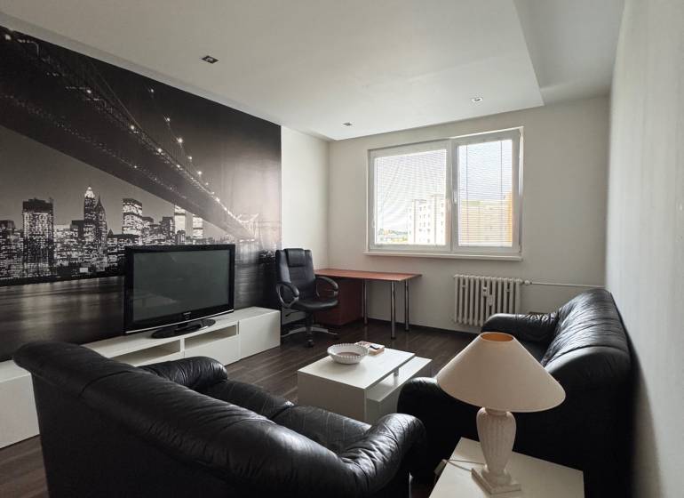 Prenájom 2-izb bytu v Bratislave-Rača - Obývacia izba s čiernou sedačkou, TV, obrazom mesta a pracovným stolom v 2-izbovom byte.