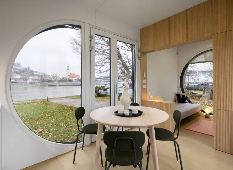 I Predaj Ecocapsule – autonómne ekologické bývanie - Kruhové okná v interiéri s výhľadom na rieku a architektúru Zvláštnych nehnuteľností.