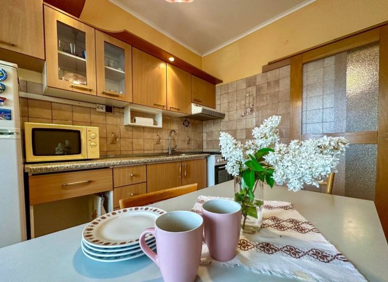 Predaj 3 - izbového bytu - Nová Baňa - Kuchyňa v 3-izbovom byte s drevenými skrinkami, kvetmi a riadom na stole.