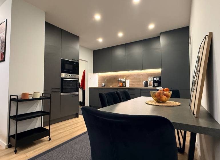 KOMPLETNE prerobený 4i byt s balkónom, 77m2 - ZÁTVOR - Kuchyňa v 4-izbovom byte s podlahou s dreveným dekorom, čiernymi skriňami a jedálenským stolom.