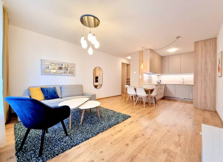 REZERVOVANÉ I Posledný investičný krásny 2-izbový byt | 62 m² | - Obývacia izba 2-izbového bytu s pohovkou, kreslom, kuchynským kútom a podlahou s dreveným dekorom.
