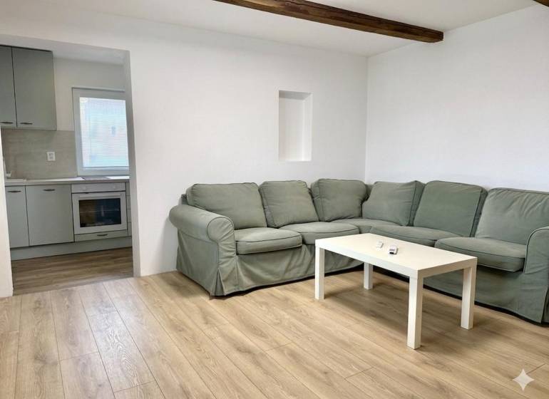 Na predaj 2 izbový murovaný domček, Bratislava, Záhorská Bystrica - Obývacia izba s rohovou pohovkou, podlahou s dreveným dekorom a kuchyňou v rodinnom dome.