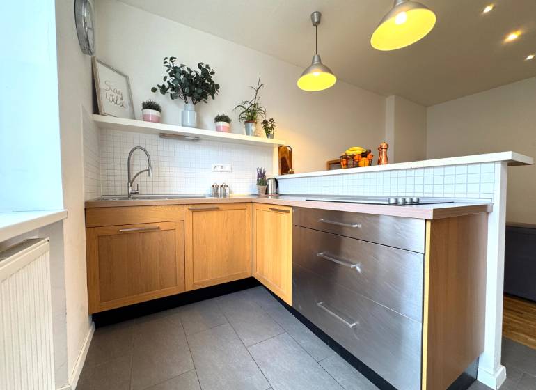 Tichý 2i byt v TOP Lokalite Nív | Trenčianska, BA - Kuchyňa v 2-izbovom byte, sivé dlaždice, drevené skrinky, závesné svetlá, rastliny na poličke.