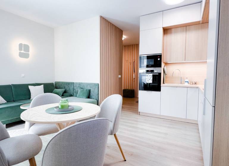 Na predaj 3 izbový byt s terasou, Trenčín, ul. Saratovská - Obývacia izba a kuchyňa v 3-izbovom byte s podlahou s dreveným dekorom a zeleným gaučom.