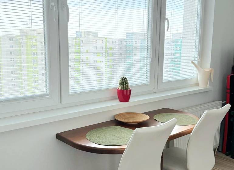 - PRENÁJOM - 1 IZBOVÝ BYT - TOPOĽČIANSKA UL. - PETRŽALKA - Stolík so židlemi pri okne v 1-izbovom byte s podlahou s dreveným dekorom.