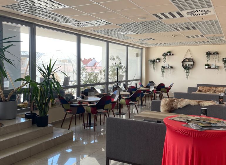 ODSTÚPENIE ZABEHNUTEJ KAVIARNE V CENTRE TRNAVY – TROJIČNÉ NÁMESTIE - Interiér gastro priestorov s kvetmi, farebnými stoličkami a pohovkami na podlahe s dekorom.