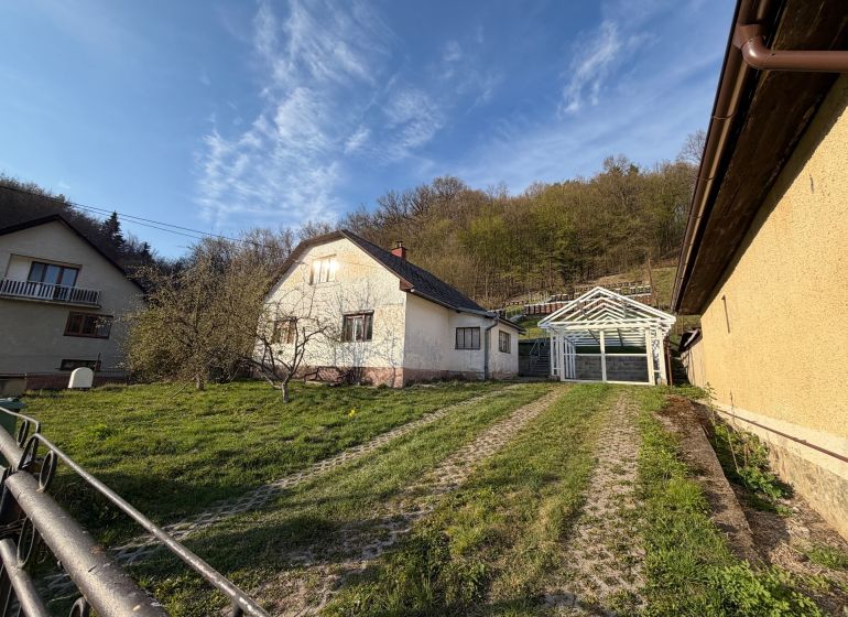RODINNÝ DOM V PRÍRODE - NA PREDAJ -OBEC ŠTÓS  - Rodinný dom v Štóse obklopený zeleňou a drevenou konštrukciou prístrešku.