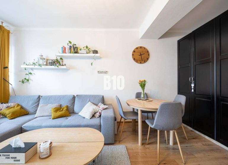 2-izbový byt po rekonštrukcii | Vietnamská ulica, Ružinov | Loggia | B - Obývačka so sivou sedačkou, okrúhlym stolom a podlahou s dreveným dekorom v 2-izbovom byte.