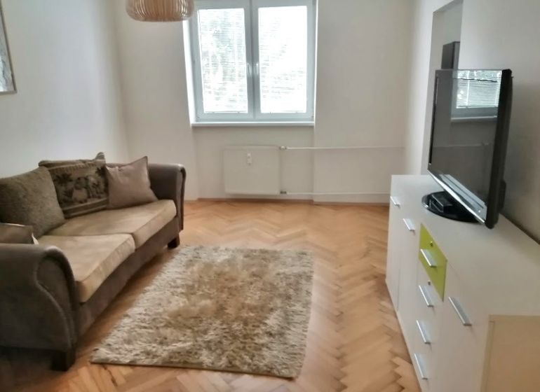 PRENÁJOM: ÚTULNÝ 2 IZBOVÝ BYT S BALKÓNOM, UHLISKO - Obývacia izba v 2-izbovom byte s pohodlnou sedačkou, televízorom a podlahou s dreveným dekorom.
