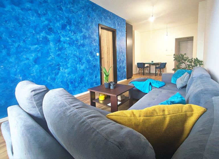 | Srdce Mesta | 2 Izb. byt | ORBIS |  70 m2 - Obývačka v 2-izbovom byte, sivý gauč, modrá stena, drevený dekor podlahy, jedálenský kút.