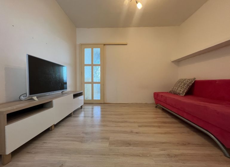 Na predaj 1-izbový byt s loggiou - Žiar nad Hronom, M. Chrasteka 1B718 - Obývacia izba s TV, červenou sedačkou a podlahou s dreveným dekorom v 1-izbovom byte.