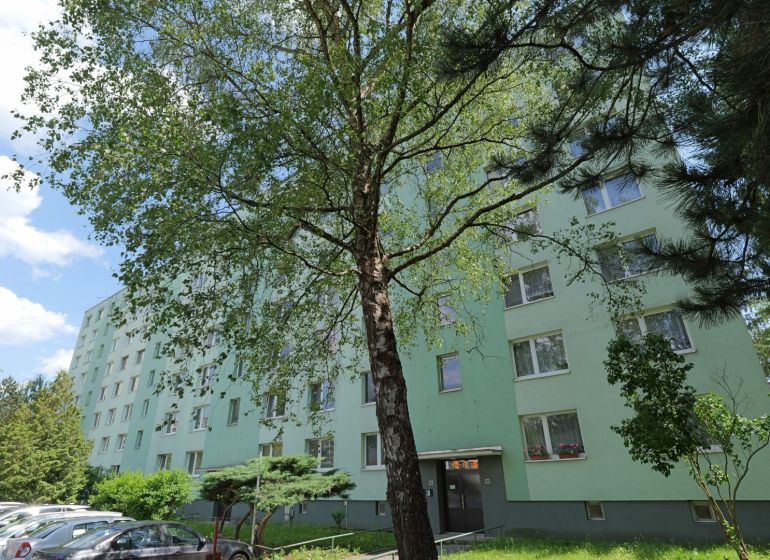 Na predaj - 3i BYT s veľkorysou výmerou - centrum PB - Považská Bystrica, Štúrova: Panelový dom obklopený stromami a zaparkovanými autami na ulici.