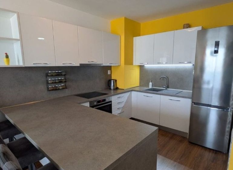 Moderný 2-izbový byt s balkónom a výhľadom - Kuchyňa v 2-izbovom byte s bielymi skrinkami, žltou stenou a podlahou s dreveným dekorom.