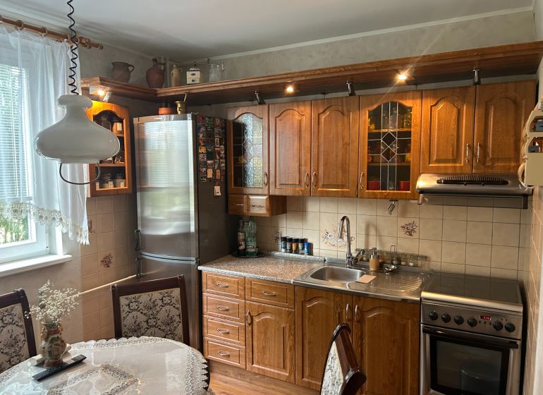 NA PREDAJ PRIESTRANNÝ 3-IZBOVÝ BYT | 67 m² | LODŽIA | 4. POSCHODIE - Kuchyňa v 3-izbovom byte s dreveným nábytkom, dekoratívnou záclonou a jedálenským stolom.