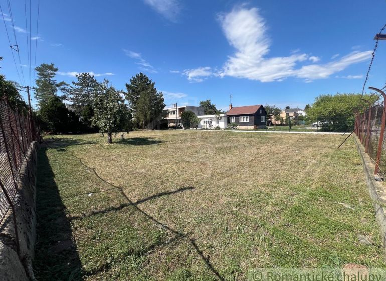 Veľký pozemok na výstavbu v centre Bernolákova. - Rovinatý pozemok v Bernolákove s trávnatou plochou obklopený stromami a oplotením.