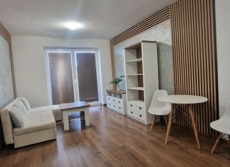 2-IZBOVÝ BYT S VEĽKÝM BALKÓNOM NA PREDAJ - Obývacia izba v 2-izbovom byte so svetlou sedačkou a podlahou s dreveným dekorom.